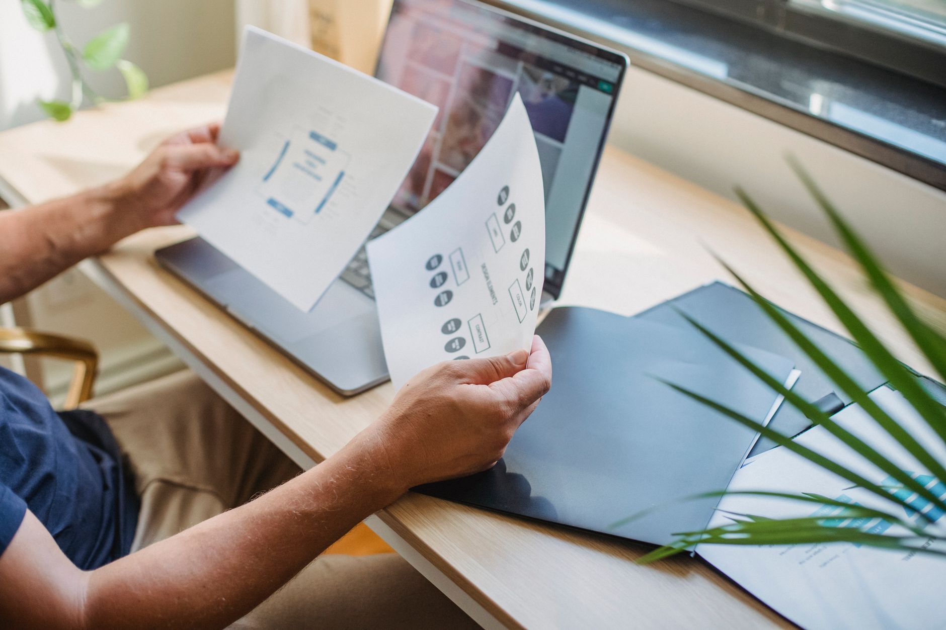 Een ontwerper zit met een laptop en mappen met papieren aan tafel