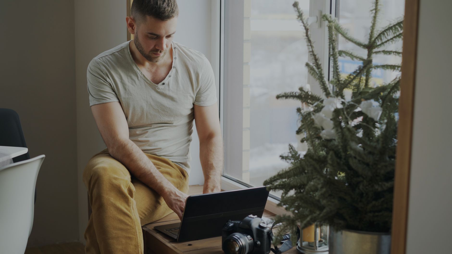 Een man werkt met een laptop bij het raam, met een camera en plant ernaast