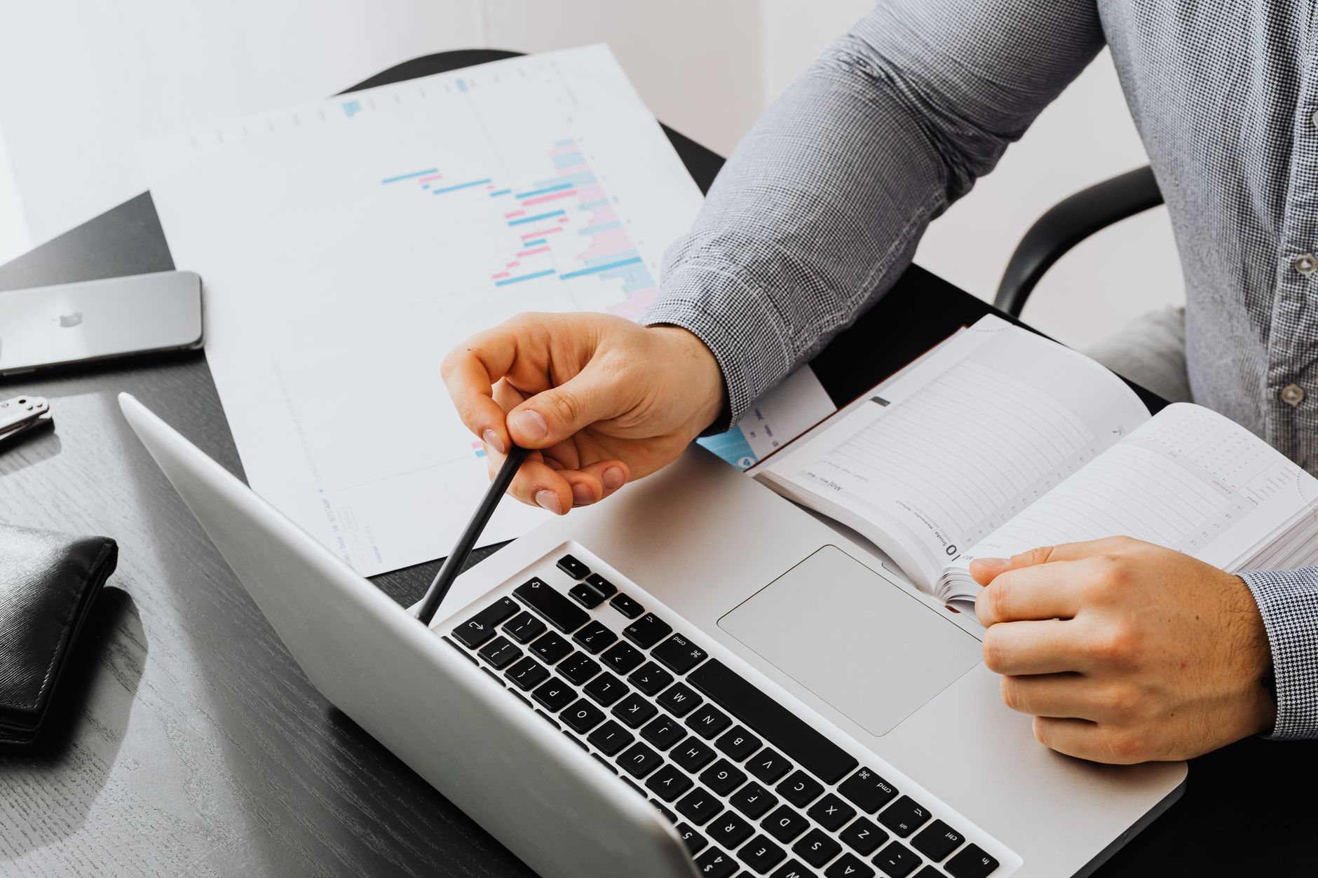 Persoon met pen bespreekt grafiek naast laptop en notitieboek op bureau