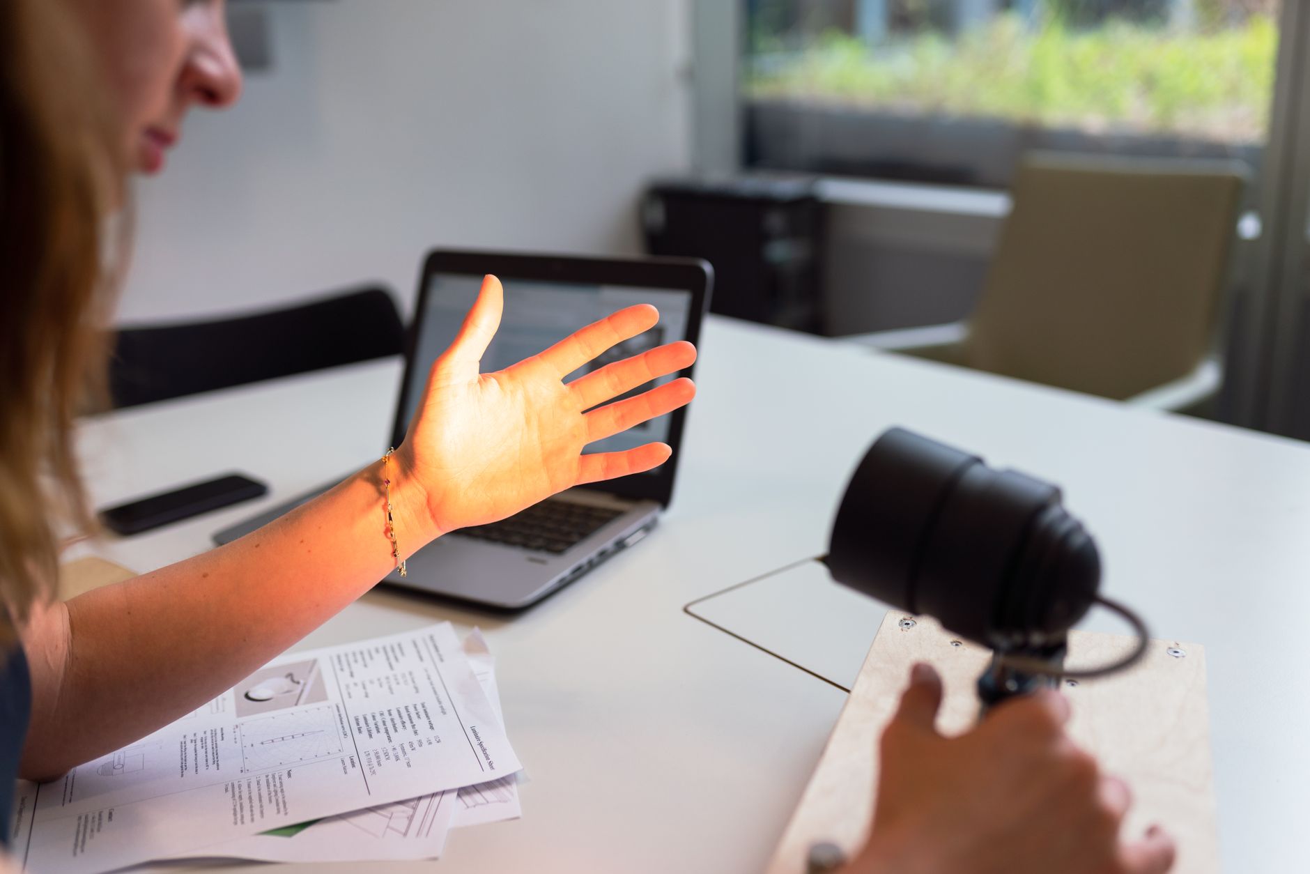 Hand gestrekt richting laptopscherm tijdens gesprek, met papieren notities op tafel